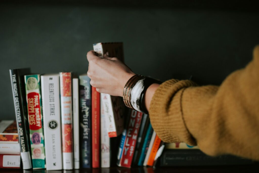 May Reading List person picking white and red book on bookshelf