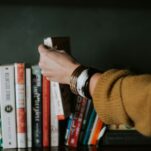 May Reading List person picking white and red book on bookshelf